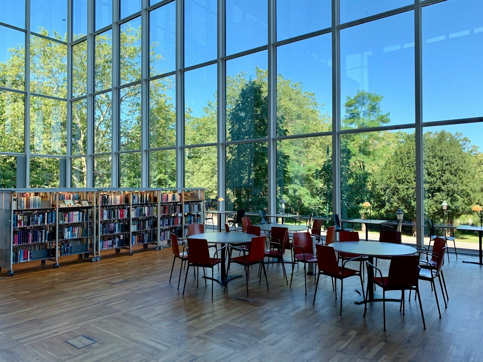 A library interior with tall windows, bookshelves, and red chairs at round tables